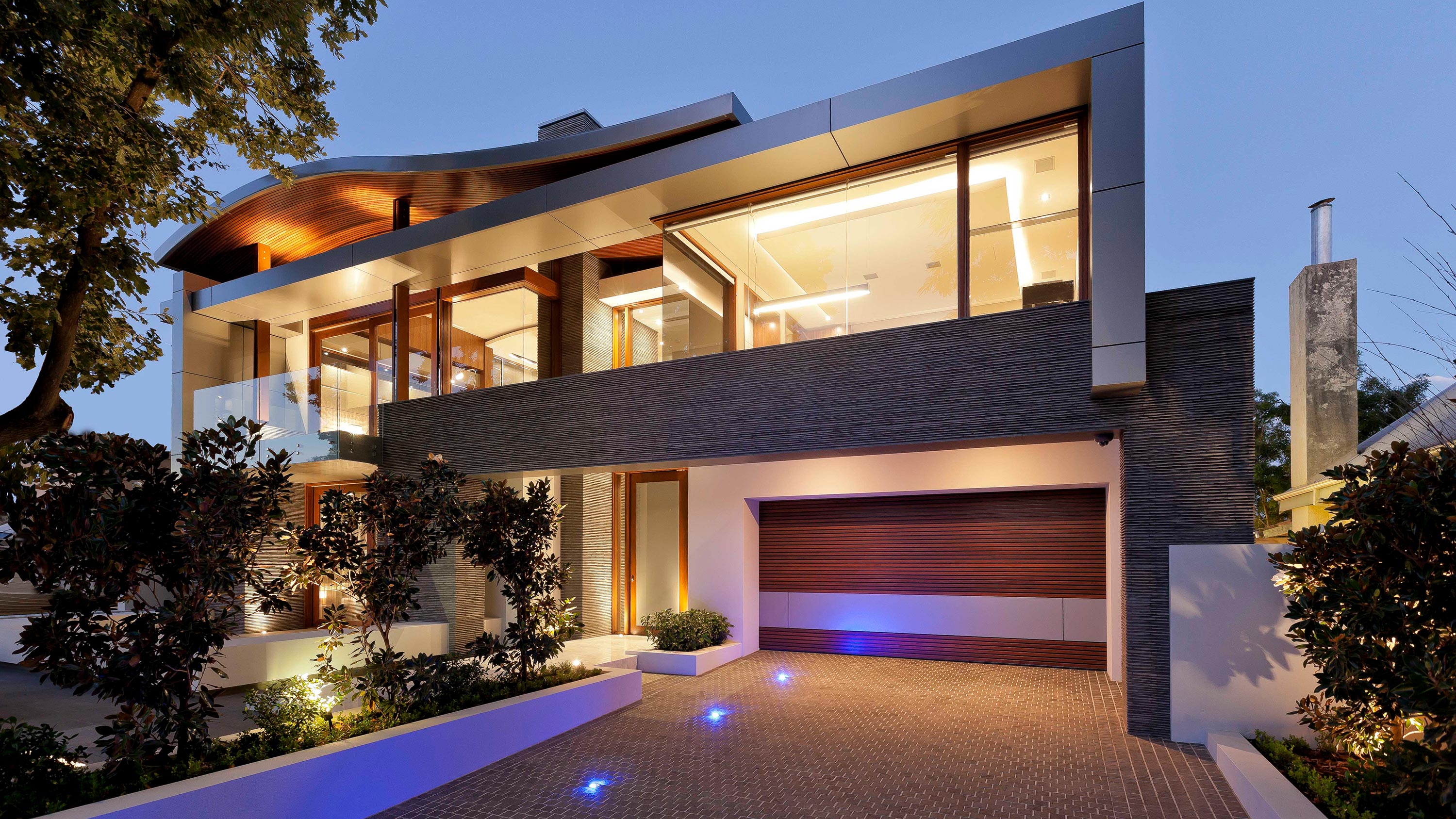 Contemporary two-story home exterior at dusk with illuminated windows, a balcony, and well-landscaped yard.