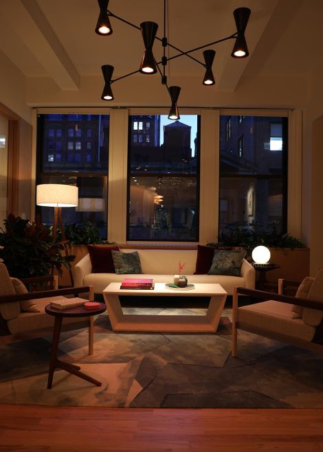 A cozy living room setup with warm lighting, a large rug, and a cityscape view through the windows.
