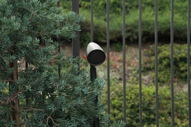 Discreet outdoor speaker mounted near greenery along a metal fence.